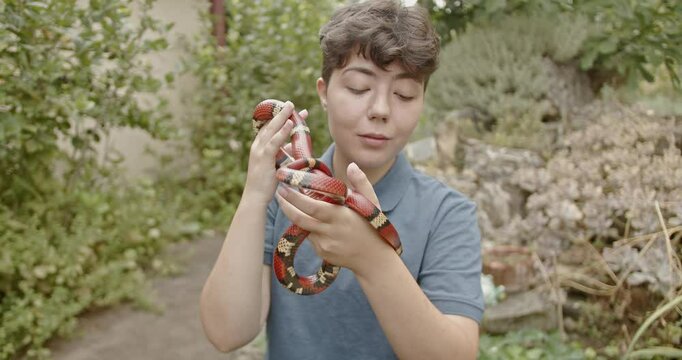 A teenage girl with short hair and a blue shirt holds her pet snake in a garden and smiles.