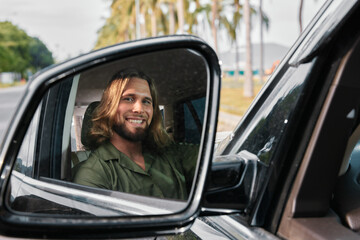A man with long hair smiles in a car s side mirror, showcasing a tropical background with palm trees and a vibrant atmosphere