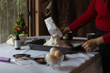 An unrecognizable brown skin woman pouring flour into a baking tray in a festive kitchen, preparing ingredients for holiday baking near a decorated Christmas tree.