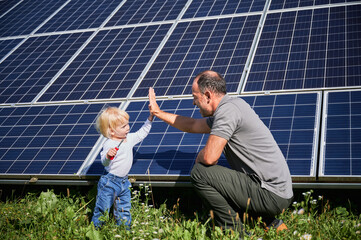 Little son and his smiling father giving each other high-five on background of solar panels. Young...