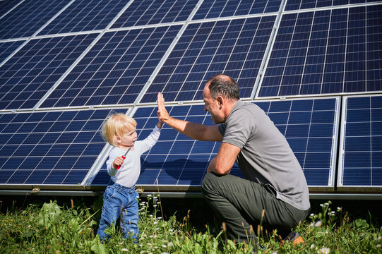 Little son and his smiling father giving each other high-five on background of solar panels. Young father glad spending time with his child. Happy family of two on background of solar panels.