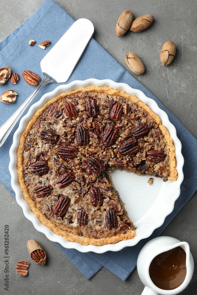 Poster Delicious pecan pie in baking dish, caramel sauce, cake server and fresh nuts on gray textured table, flat lay