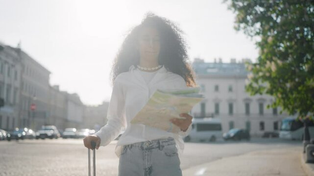 Full-length portrait of young Arabic woman with big suitcase in city center. Traveling in summer vacation, female tourist viewing paper map, sightseeing and orientation in town, slender tall lady