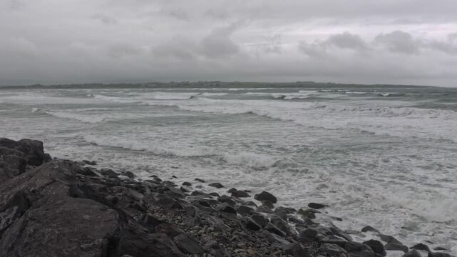 Diagonal view of Strandhill Beach, Ireland, during a storm in the Atlantic Ocean. Choppy waves crash against the rocky shoreline under dark clouds