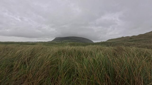 Wind sweeps through coastal dunes near Strandhill Beach, Ireland, with thick clouds hanging over the distant mountain. The movement of the grass captures the dynamic nature of this rugged landscape