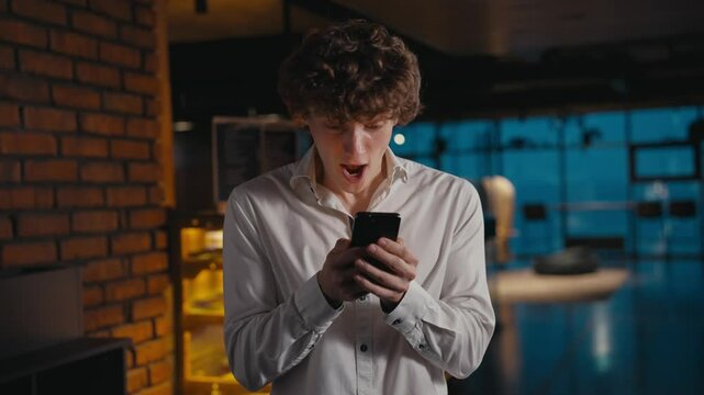 A young guy with curly hair is pleasantly surprised and jumps with happiness when he looks at the screen of his phone in the office