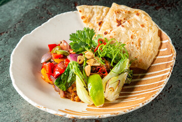 A vibrant plate of food featuring a side of flatbread, a colorful vegetable salad with mixed greens, and garnished with fresh herbs on a patterned ceramic plate against a dark background.