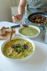 Woman serving dinner with a fresh and homemade cooked vegetarian pea soup with fried mushrooms in the kitchen