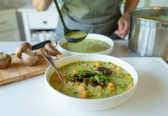Woman serving with a ladle a delicious homemade, vegetarian pea soup with mushrooms in the kitchen