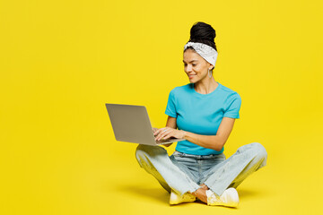 Full body young smiling happy fun IT Latin woman wears blue top t-shirt headband casual clothes sit hold use work on laptop pc computer isolated on plain yellow background studio. Lifestyle concept.