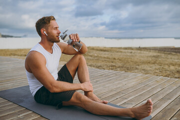 Young strong athletic toned fit sportsman man he wear sports clothes earphones hold bottle drink...