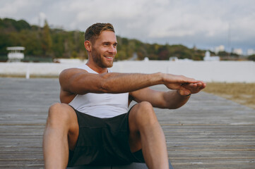 Young strong sporty fit sportsman man wears sports clothes do abdominal abs exercises at floor, pump press warm up training at sunrise sun over sea beach outdoor on pier seaside in summer day morning.