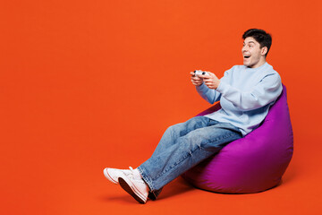 Full body young man he wearing blue sweatshirt casual clothes sit in bag chair hold in hand play pc game with joystick console isolated on plain red orange color background studio. Lifestyle concept.