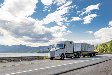 Bonnet powerful white big rig semi truck transporting covered fastened cargo on flat bed semi...