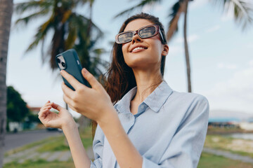 Woman wearing sunglasses, smiling at her cell phone while standing in front of palm trees,...