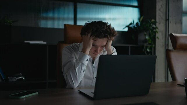 A tired guy with curly hair in a white shirt looks tiredly at a gray laptop and holds his head with his hands during a hard day at work in the office