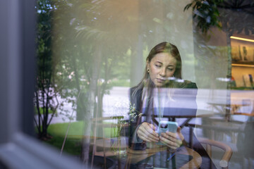 View through glass interested female scrolling news social media feed on smartphone rest in cafe with cup. Businesswoman have coffee break using mobile phone surfing internet web enjoy coffee.
