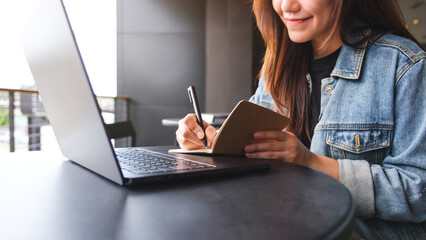 Closeup image of a young woman writing on a notebook while working on laptop computer