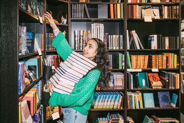 Girl with heap of books reaching at shelf while standing in bookstore