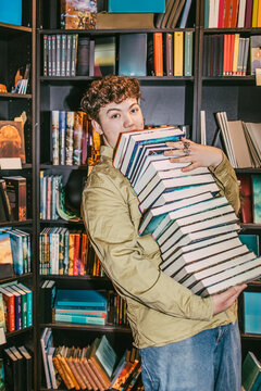 Portrait of young man balancing heap of books near shelf in bookstore