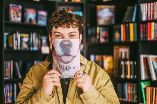Portrait of young man with curly hair covering face with book in bookstore