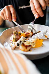  close-up of hands using a fork and knife to eat a gourmet dish on a white plate. The dish features various colorful elements, showcasing a fine dining experience. Perfect for culinary themes.