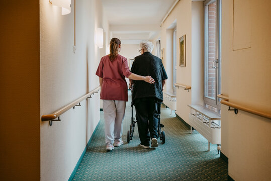 Rear view of female caregiver assisting senior woman walking with mobility walker in corridor