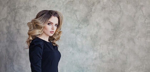 Fototapeta premium Perfect young woman with curly hair and make-up in black dress posing against gray stucco plaster background wall with copy space