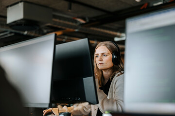 Focused female entrepreneur with wireless headphones doing coding on computer in office