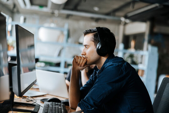 Side view of young programmer with hand on chin sitting and looking at computer in office