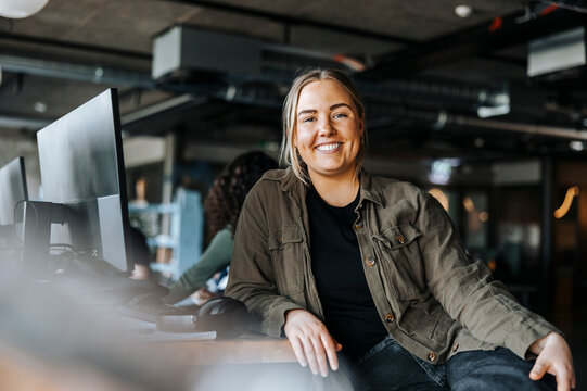 Portrait of happy female tech programmer leaning on desk sitting at office