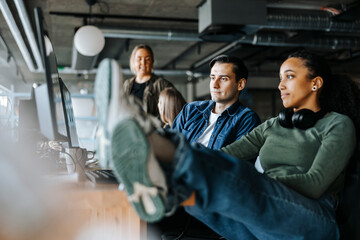 Female coder sitting with feet up sharing computer screen with male colleague while sitting in office