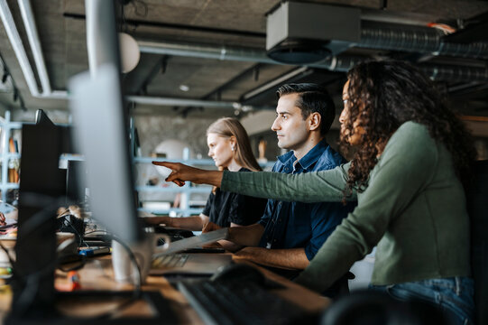 Female coder pointing at device screen while sitting with colleagues in office