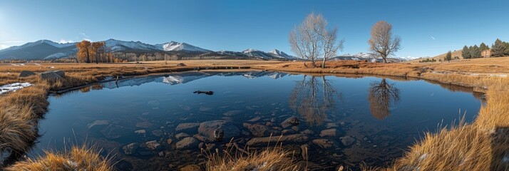 A serene autumn landscape featuring a crystal-clear pond reflecting snow-capped mountains, barren trees, and golden grasses under a clear blue sky