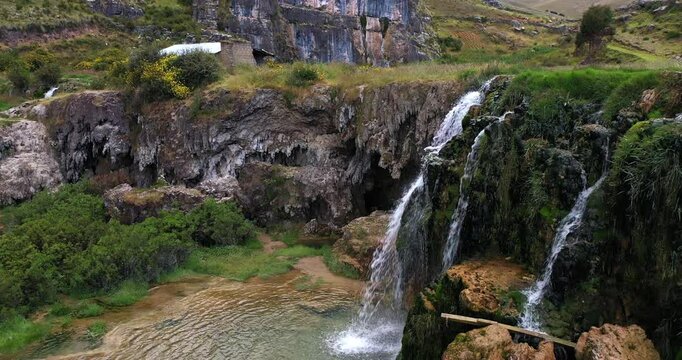 Beautiful drone shot of a natural waterfall next to the Millpu lake of turquoise waters with lots of vegetation and mountains around on a cloudy day.