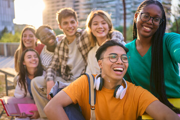 Obraz premium Happy multiracial classmates university students posing sitting together taking selfie photo outdoors. Generation z friends smiling having fun looking joyful at phone for group picture in sunny park 