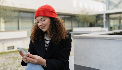 pretty curly woman in city street in autumn coat, using smart phone, red knitted hat