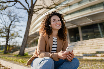 pretty curly smiling woman walking in city street in stylish jacket, using smartphone