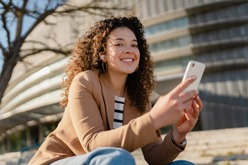 pretty curly smiling woman walking in city street in stylish jacket, using smartphone