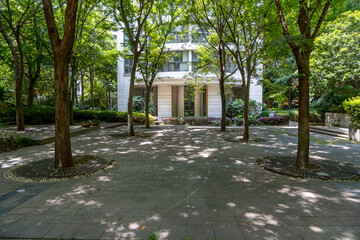 Urban outdoor green space with a paved walkway, dappled with sunlight filtering through the leaves of tall, mature trees, and a white residential apartment building in the background.