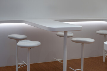 A minimalist, modern cafe or restaurant interior featuring a sleek, white counter table and stools...