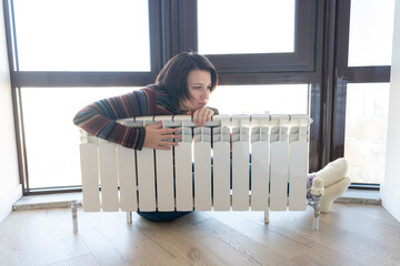 Woman wearing pullover sitting near heater radiator and hugs it