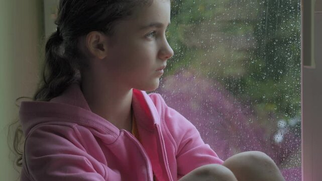 Alone Sad Little Girl Depressed by Bad Weather Watching Rain Through Window. Disappointed Child Unhappy Bored Lonely and Frustrated. Pensive Child Looking out Window During Rainy Day. Sadness Sorrow.