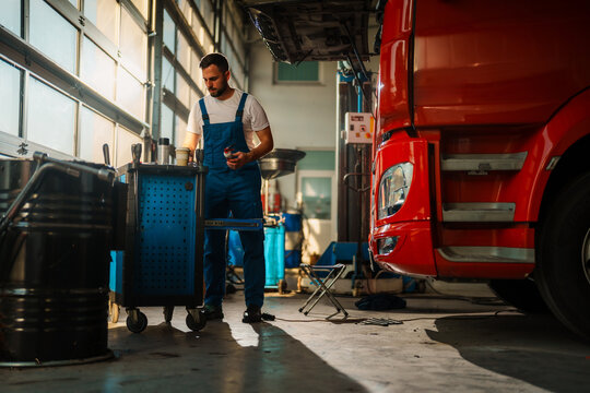 Mechanic in blue working suit work on a truck in a garage. The scene is well-lit with sunlight streaming through large windows, highlighting the tools and equipment around.