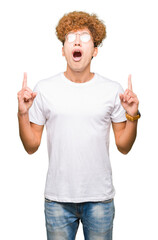 Young handsome man with afro hair wearing glasses amazed and surprised looking up and pointing with fingers and raised arms.