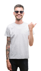 Young tattooed adult man wearing sunglasses pointing and showing with thumb up to the side with happy face smiling