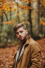 A man in a brown jacket is standing in a forest with leaves on the ground
