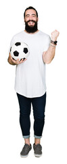 Young man with long hair and bear holding soccer football ball screaming proud and celebrating victory and success very excited, cheering emotion