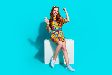 Full size photo of pretty young girl sit cube hold device winning wear trendy colorful outfit isolated on aquamarine color background