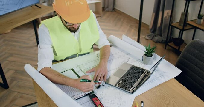 Top view. Man designer in helmet and vest which working with architectural scheme on blueprint in workroom